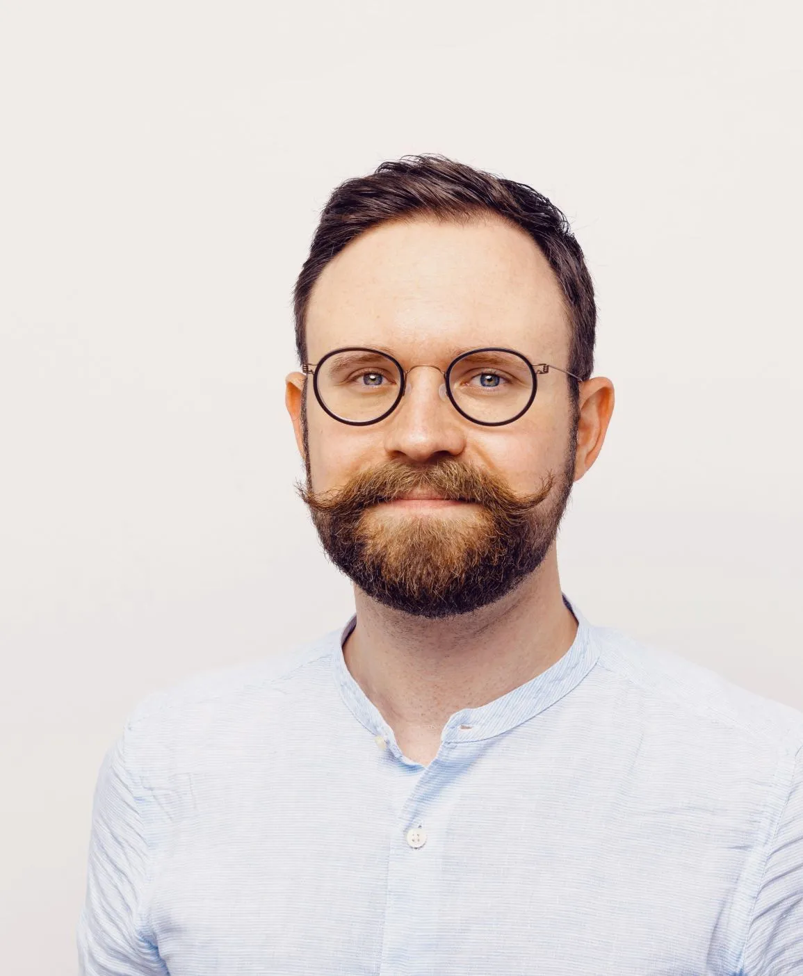 Portrait of Florian Pichler, wearing black rimmed glasses, mustache and beard, in a light blue shirt
